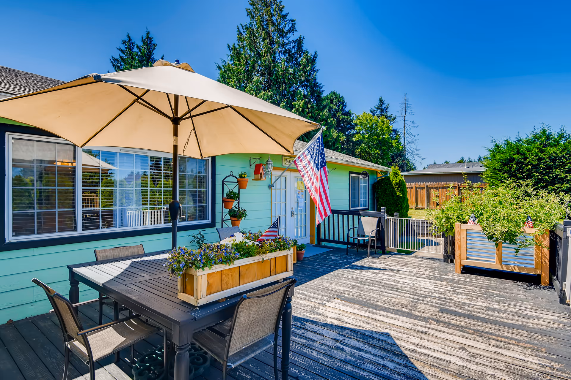 Sunny outdoor wooden deck with a dining table and umbrella, planter boxes, American flags, and a mint-green building entrance.
