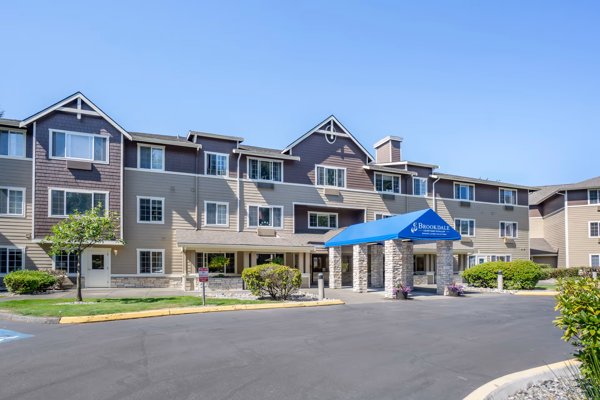 Exterior view of Brookdale Courtyard Puyallup senior living facility showing a three-story building with beige and brown siding, multiple windows, a blue canopy entrance supported by stone pillars, and surrounding landscaping with bushes and a small tree under a clear blue sky.