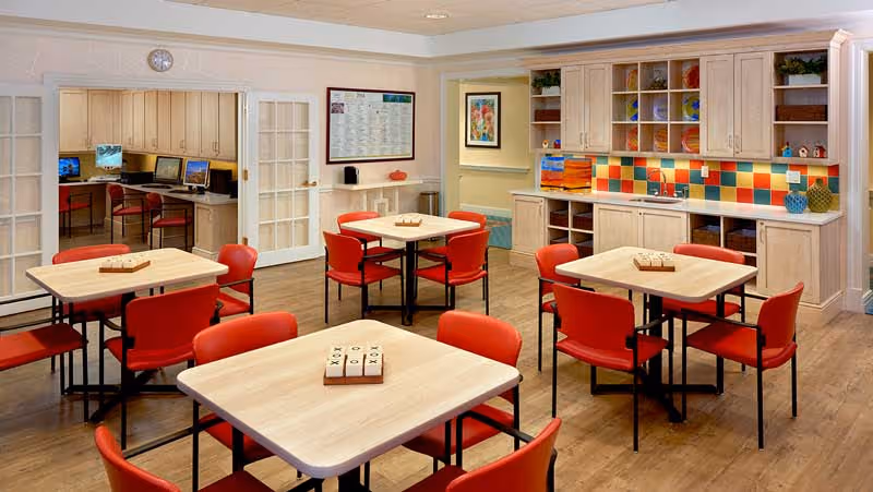 A bright and clean common area with several square tables surrounded by red chairs. Each table has a tic-tac-toe game set on it. The room features a kitchenette with colorful tiled backsplash, light wood cabinets, and a countertop. In the background, there is a small computer area with multiple desktop computers and chairs. The walls are decorated with framed artwork and a bulletin board.