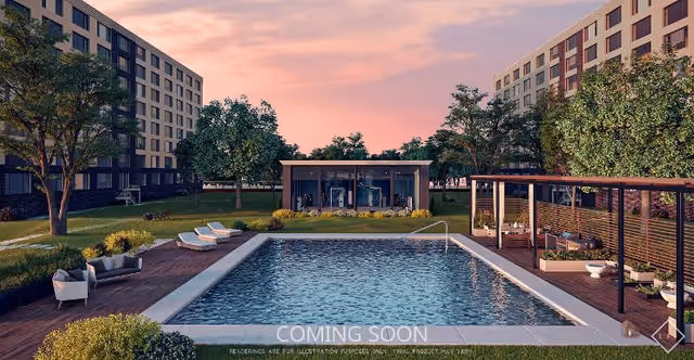 Outdoor view of a senior living facility featuring a rectangular swimming pool surrounded by lounge chairs and seating areas. The pool area is bordered by wooden decking and greenery, with two multi-story buildings in the background and a small glass-enclosed structure near the pool. The sky is at sunset with a pinkish hue.