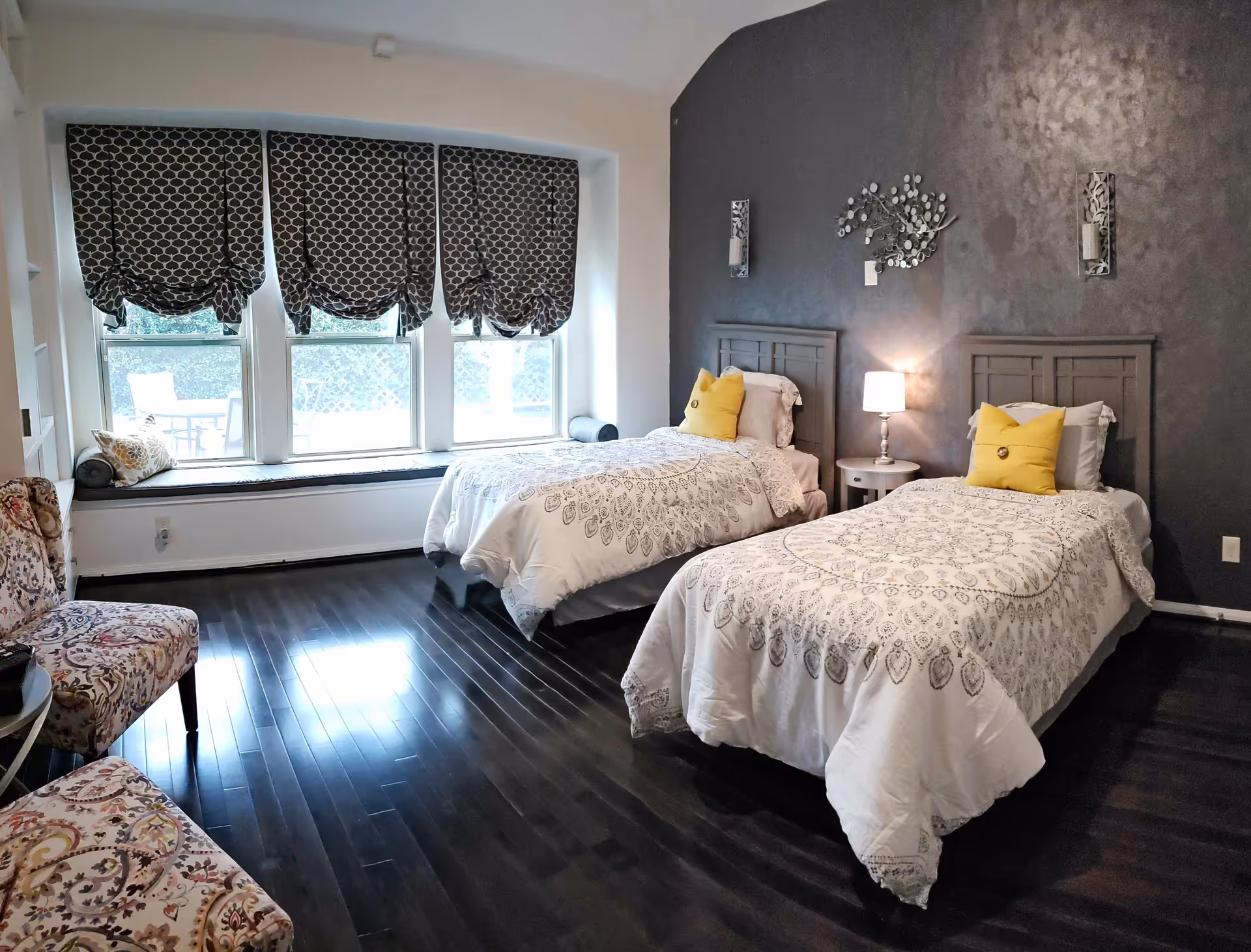A bedroom with two twin beds featuring white and gray patterned bedding and yellow accent pillows. The room has dark hardwood floors, a dark gray accent wall with decorative wall art and sconces, a small round bedside table with a lamp, three large windows with patterned Roman shades, and a cushioned window seat. There are also two patterned chairs and a small round table in the corner.