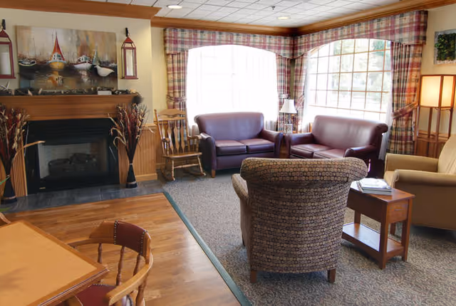 A cozy living room area with two purple leather sofas, a patterned armchair, a wooden rocking chair, and a beige armchair. There is a fireplace with a wooden mantel decorated with a painting of boats and two red lanterns. Large windows with plaid curtains allow natural light to fill the room. A wooden side table with a lamp and a small table with chairs are also visible.