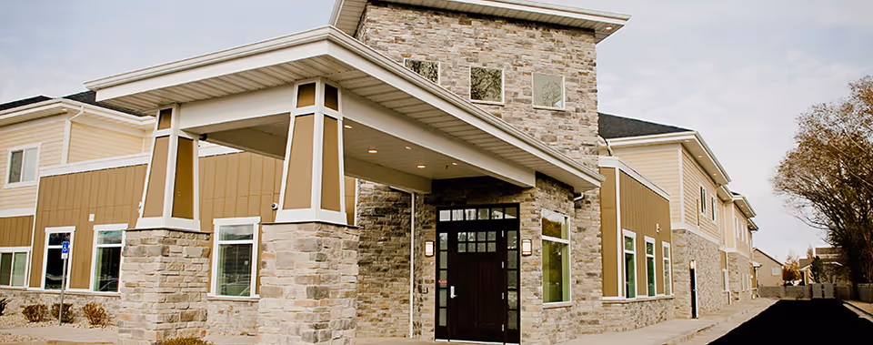 Exterior view of a modern assisted living facility building with stone and beige siding, large windows, and a covered entrance supported by stone pillars. The sky is partly cloudy and there are some trees visible on the right side.