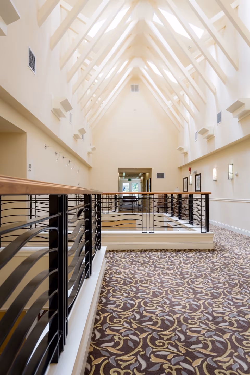 Interior hallway with a high vaulted ceiling featuring exposed beams and skylights. The floor is covered with a patterned carpet in shades of brown, beige, and gray. Black metal railings with wooden handrails line the walkway, and framed artwork and wall sconces are visible on the right wall. The hallway leads to a doorway with glass panels at the far end.