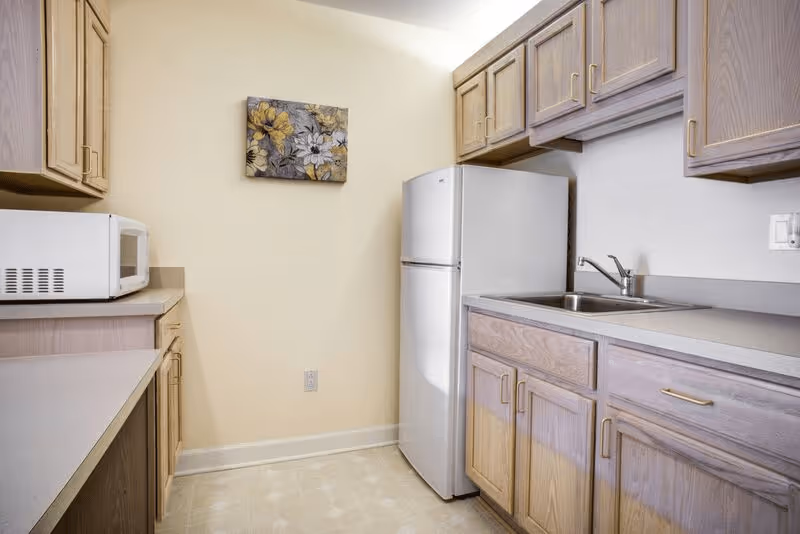 Small kitchen area with light wood cabinets, a white refrigerator, a microwave on the countertop, a stainless steel sink, and a floral painting on the wall.