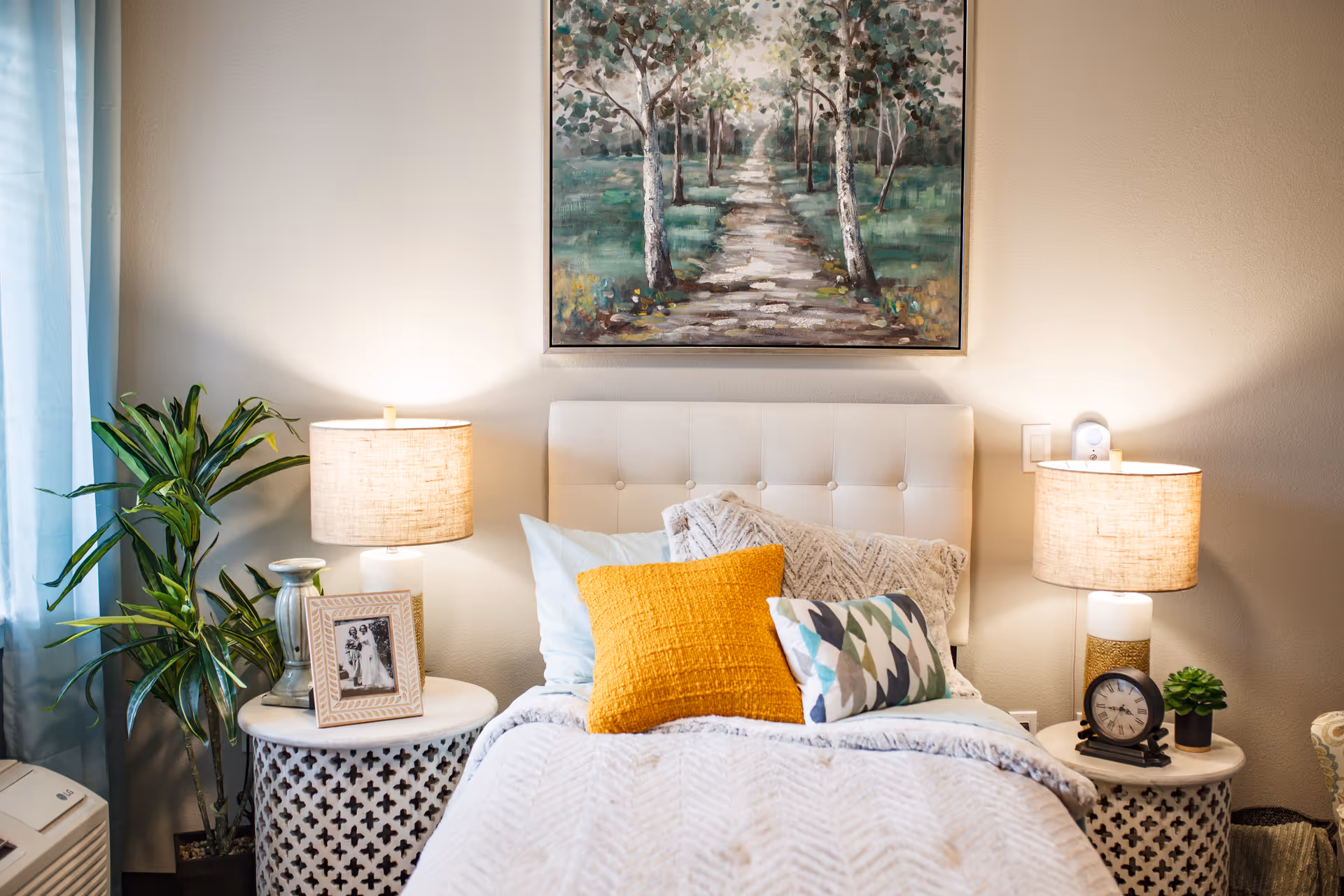 A cozy bedroom with a white tufted headboard, a bed with white and patterned pillows including a yellow one, and a textured white blanket. On either side of the bed are round white nightstands with decorative cutouts, each holding a lamp with a beige shade. The left nightstand also has a framed black and white photo and a small vase, while the right nightstand has a clock and a small green plant. A large painting of a tree-lined path hangs above the bed, and a tall green plant is placed near the window with blue curtains on the left side.