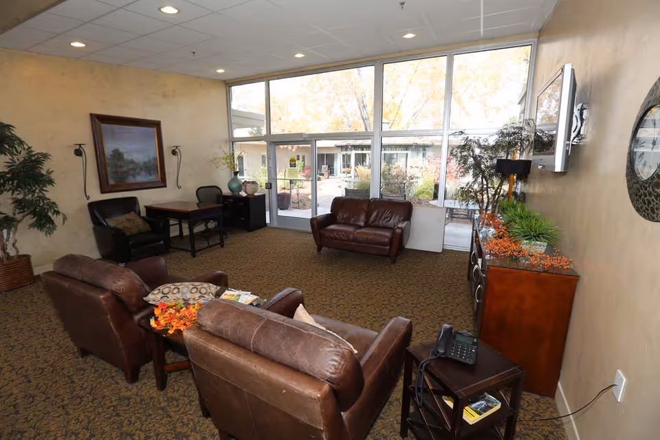 A furnished lounge with leather chairs and sofas, a TV and side tables, and large windows overlooking an outdoor courtyard.