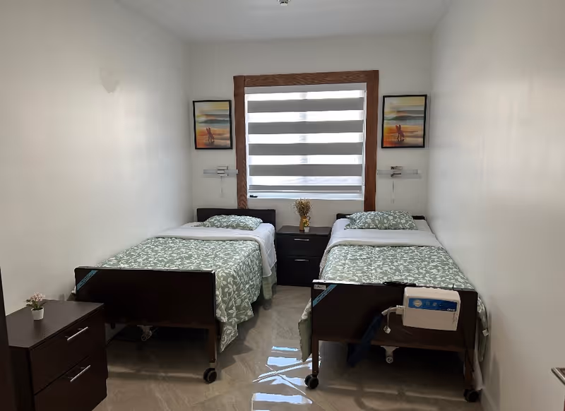 A small, clean bedroom with two single beds placed parallel to each other with a nightstand in between. Each bed has green and white patterned bedding and a pillow. There are two framed pictures on the wall, one above each bed, and a window with a striped blind in the center. A small drawer unit with a potted plant is on the left side of the room. The floor is tiled and the walls are white.
