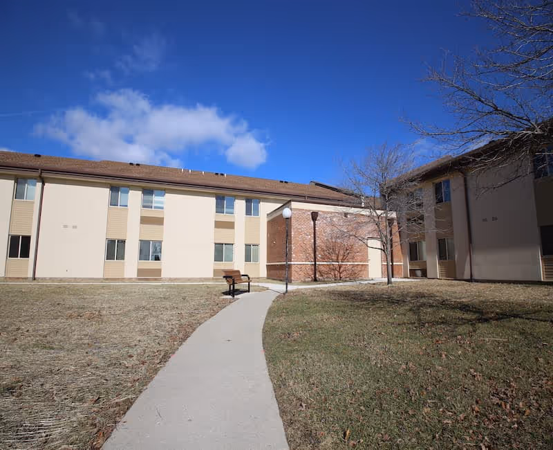 Outdoor view of a senior living facility with a paved walkway leading to a bench and two buildings with beige and brown exterior walls under a blue sky with some clouds.