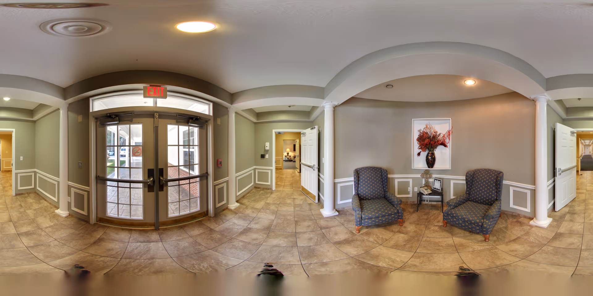 Interior view of a senior living facility entrance area with double glass doors marked with an exit sign. The space features tiled flooring, light green walls with white trim, two patterned armchairs, a small table with decorative items, and a framed floral artwork on the wall. There are doorways leading to other rooms on either side of the entrance.