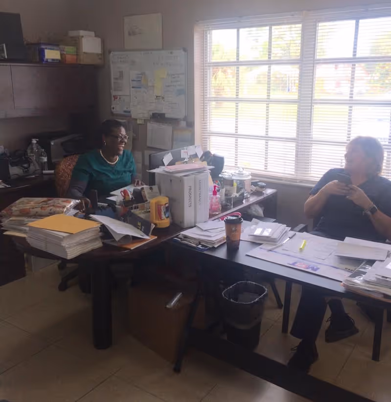 Two women sitting at desks in an office filled with papers, folders, and office supplies. One woman is smiling and looking at the other, who is holding a phone. A large window with blinds lets in natural light behind them.