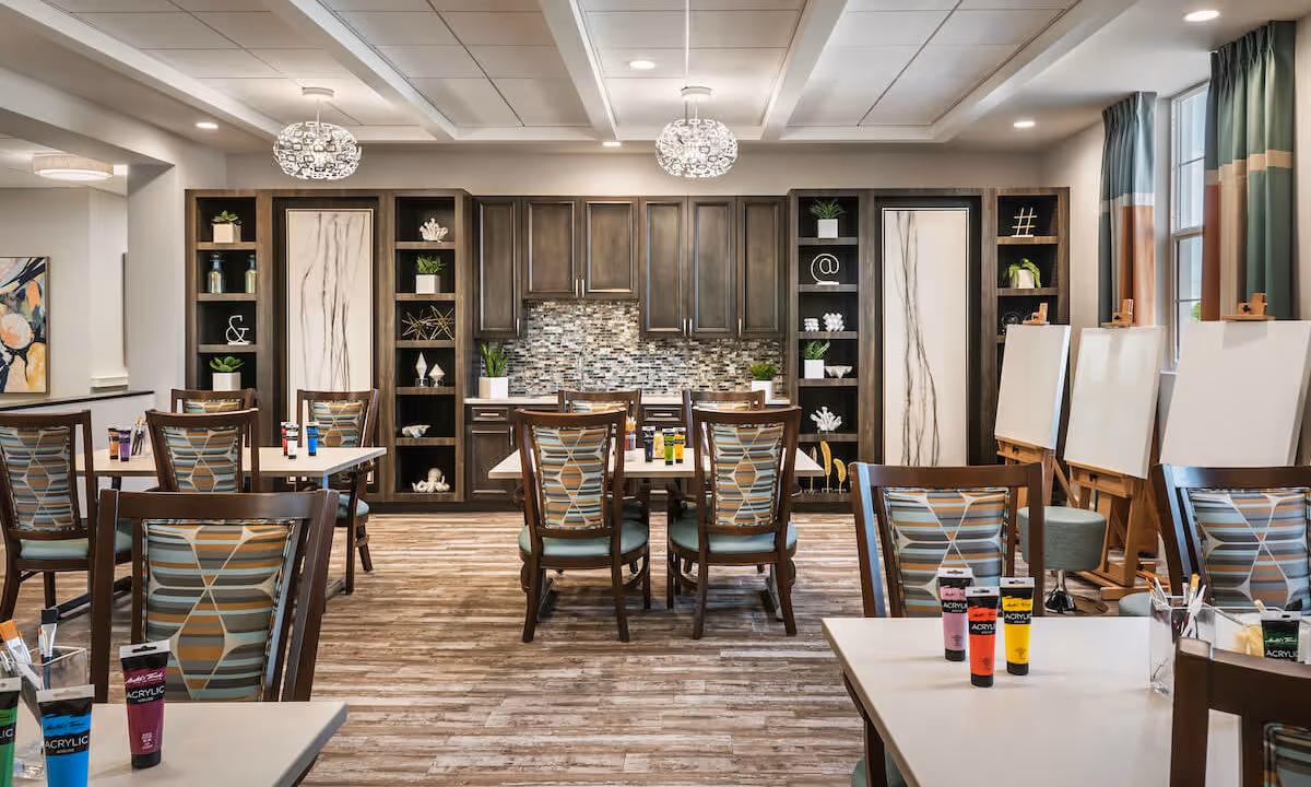 A bright and spacious art activity room in a senior living facility with multiple tables and chairs arranged for painting. The tables have acrylic paint tubes and paintbrushes. The back wall features dark wooden cabinets with decorative shelves and a mosaic tile backsplash. Large windows with green and beige curtains let in natural light, and there are several easels with blank canvases set up near the windows.