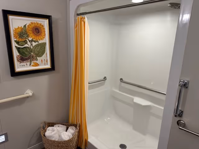 A clean, accessible shower area with white walls, a built-in seat, and metal grab bars on the walls. A yellow shower curtain is partially drawn to the side. On the left wall, there is a framed picture of sunflowers and a basket with rolled white towels underneath it.