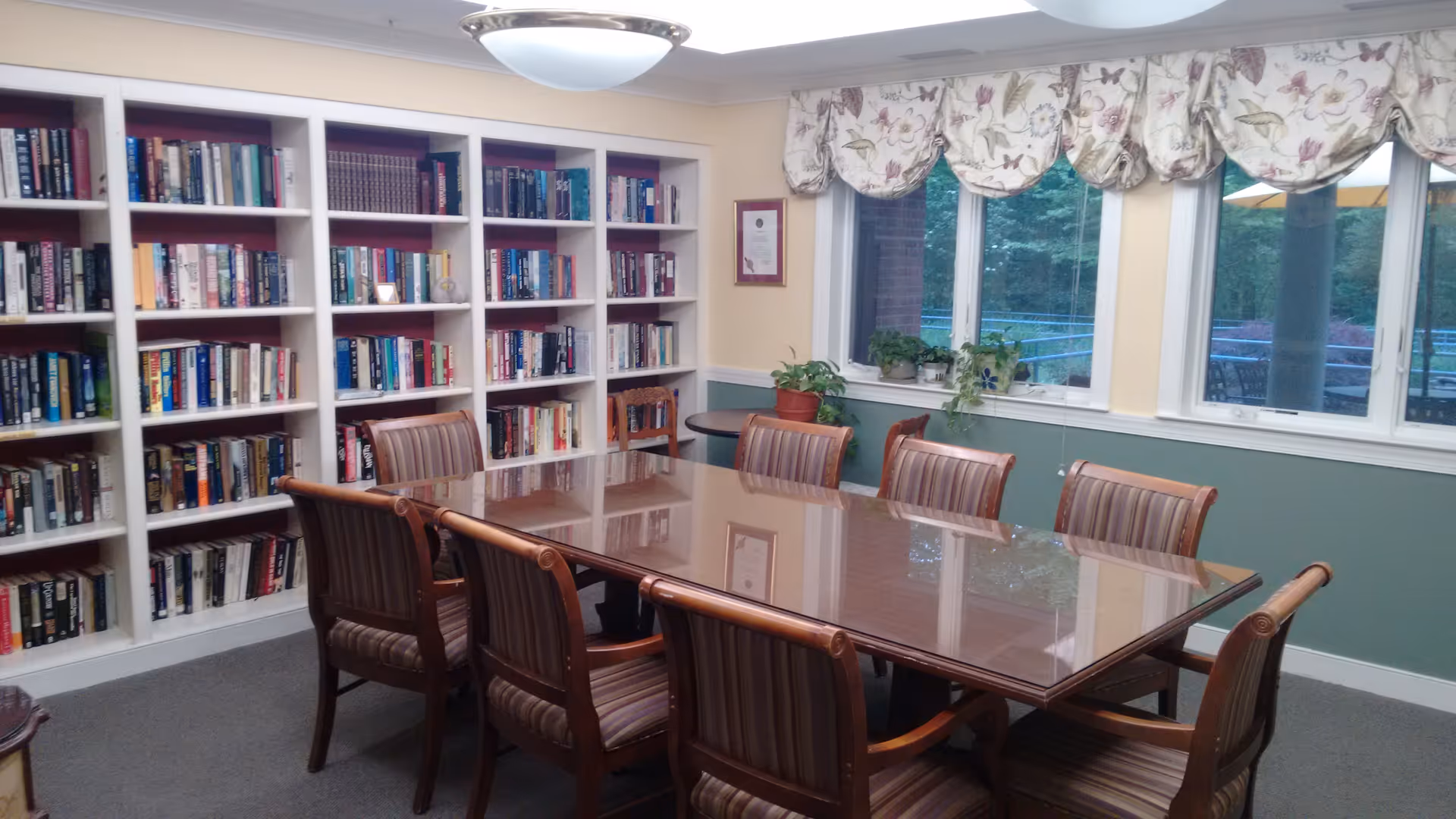 A communal room with a large polished wooden table and chairs, bookshelves along one wall, and windows with floral valances.
