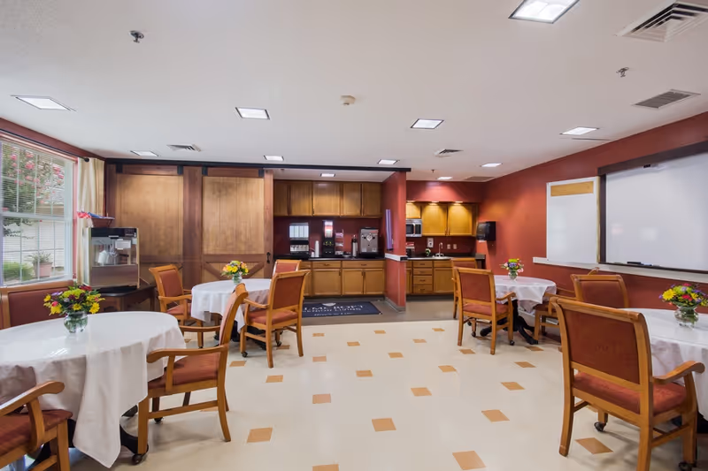 A bright and clean dining room with round tables covered in white tablecloths, each adorned with a small vase of colorful flowers. Wooden chairs with red cushions surround the tables. The room features a kitchenette area with wooden cabinets, coffee machines, and a sink. Large windows on the left side let in natural light, and the walls are painted a warm red color. There is a whiteboard on the right wall.