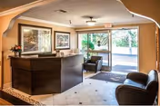 Reception area of Palm Terrace Care Center featuring a dark wooden front desk with a vase of flowers on the left, two framed landscape paintings on the wall behind the desk, a tiled floor, and a glass door entrance with a welcome mat. There are two dark leather chairs positioned near the entrance.