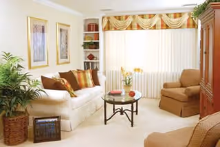 Bright, neatly furnished living room with a sofa, armchair, coffee table, plants, and a large window with drapes.