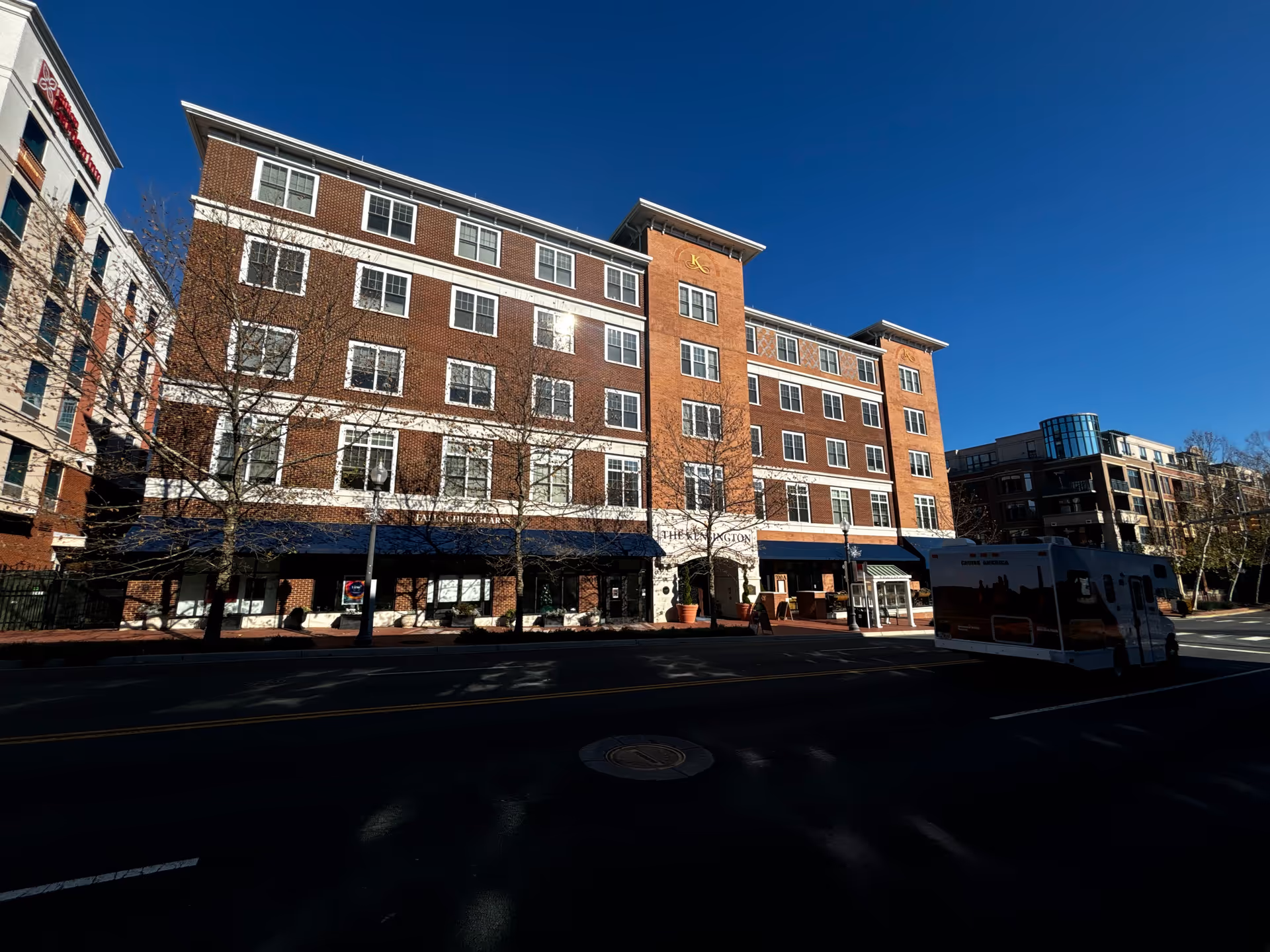 Exterior view of The Kensington Falls Church building, a multi-story brick and stone structure with multiple windows, a blue awning over the entrance, and a clear blue sky above. A vehicle is parked on the street in front of the building.