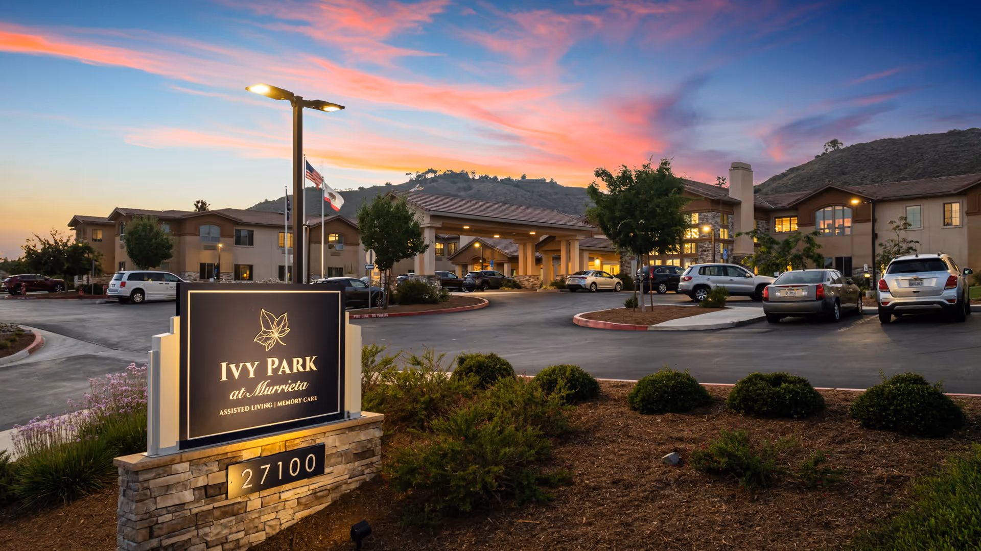 Exterior view of Ivy Park at Murrieta assisted living and memory care facility at sunset, showing the building, parking lot with cars, landscaped bushes, and a sign with the facility name and address 27100.