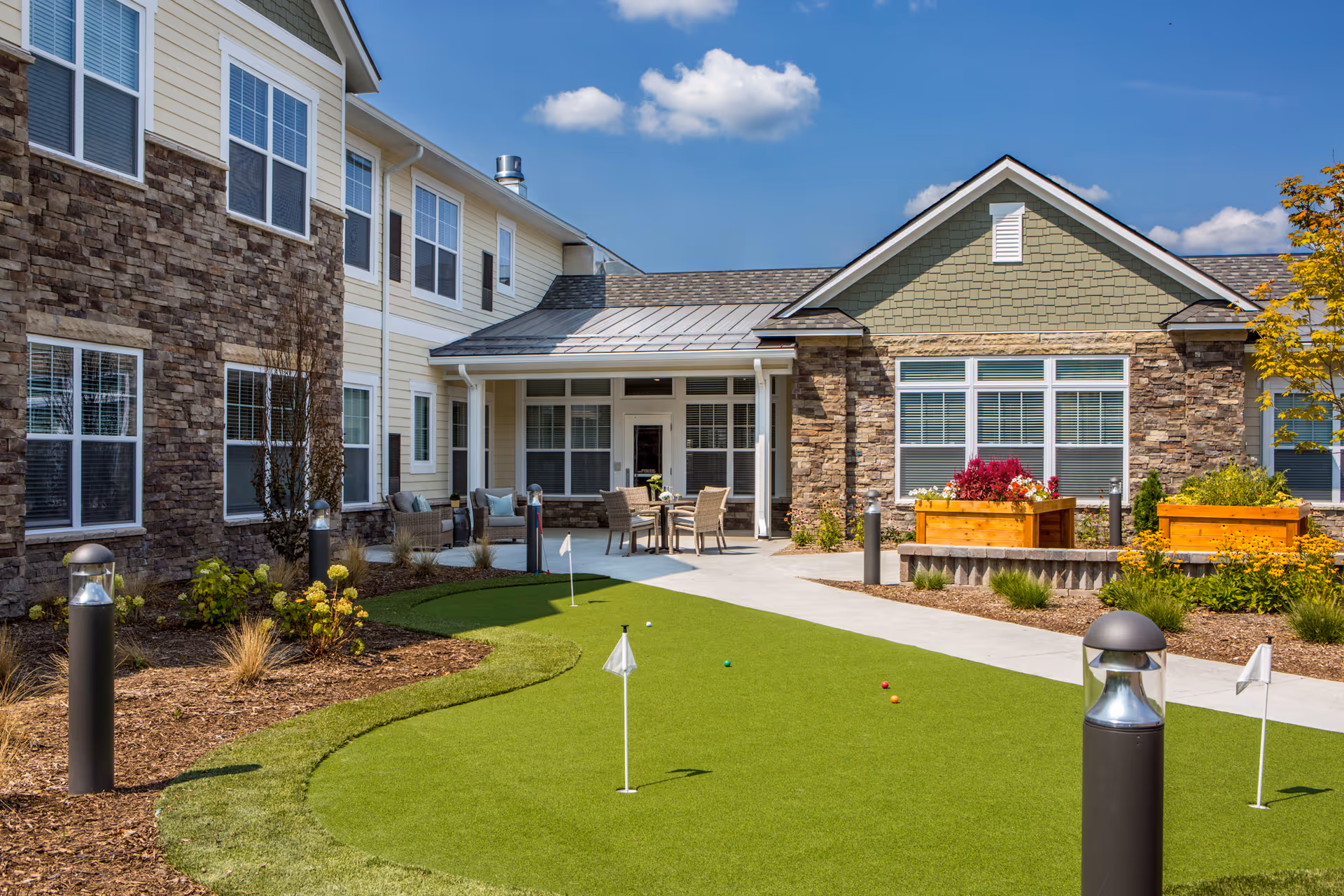 Outdoor courtyard area of a senior living facility with a putting green, patio seating with chairs and tables, landscaped garden beds with flowers and shrubs, and a two-story building with stone and siding exterior under a blue sky with some clouds.
