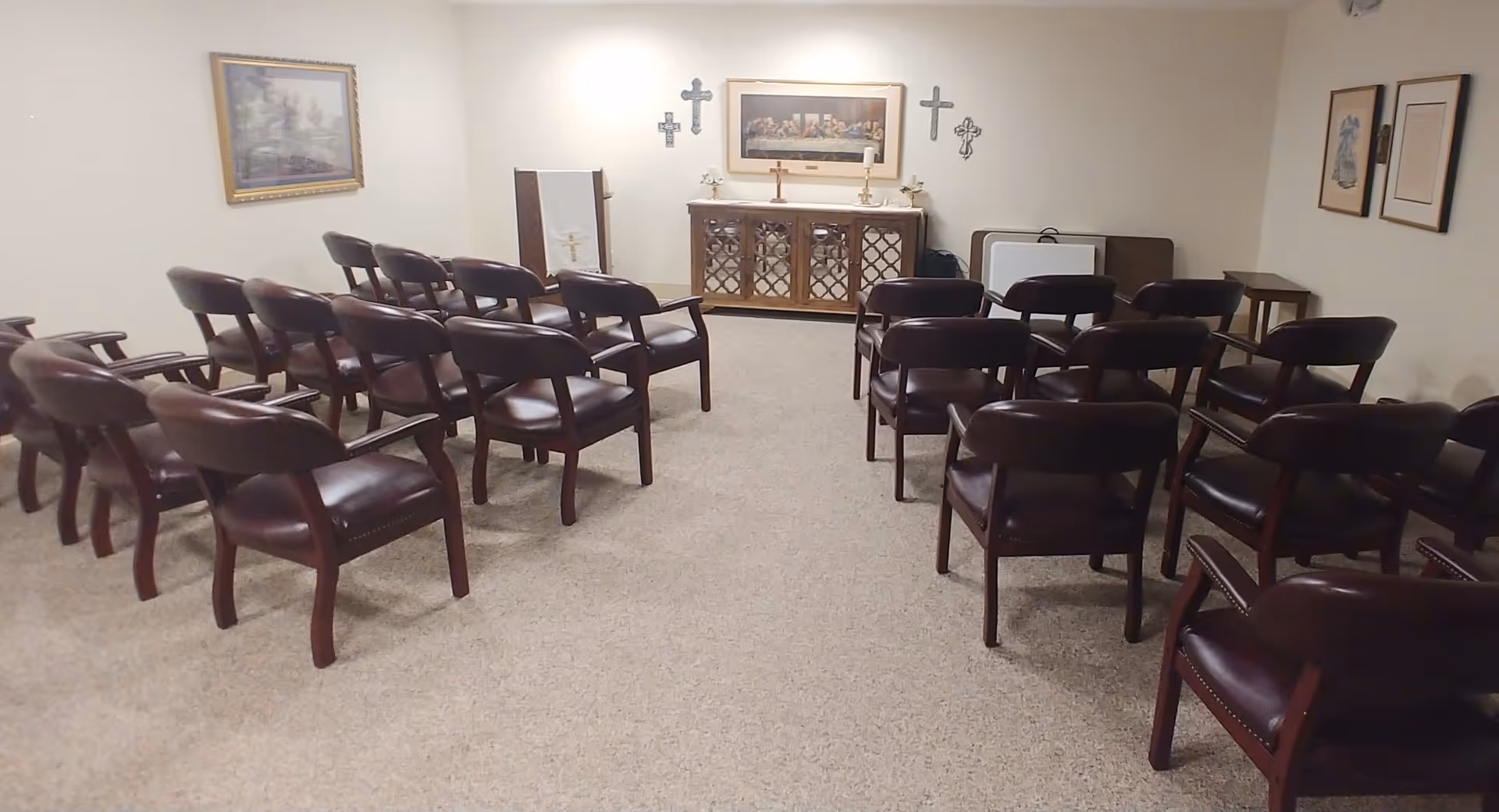 A small chapel or prayer room with rows of brown wooden chairs with armrests facing a cabinet adorned with religious crosses and a framed picture above it. The room has beige carpet and light-colored walls with framed artwork.