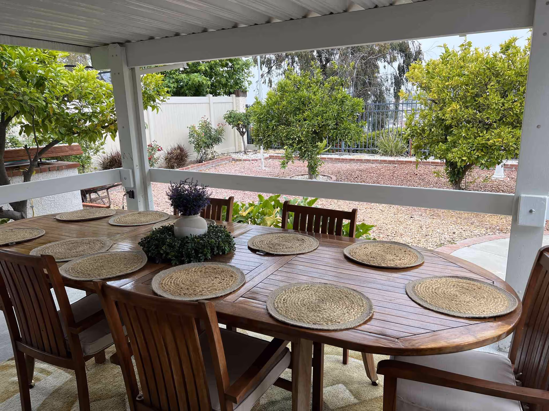 Outdoor covered patio area with a large wooden dining table set with eight woven placemats and a centerpiece of a white vase with purple flowers surrounded by greenery. The patio overlooks a landscaped garden with small trees, bushes, and a gravel ground cover.