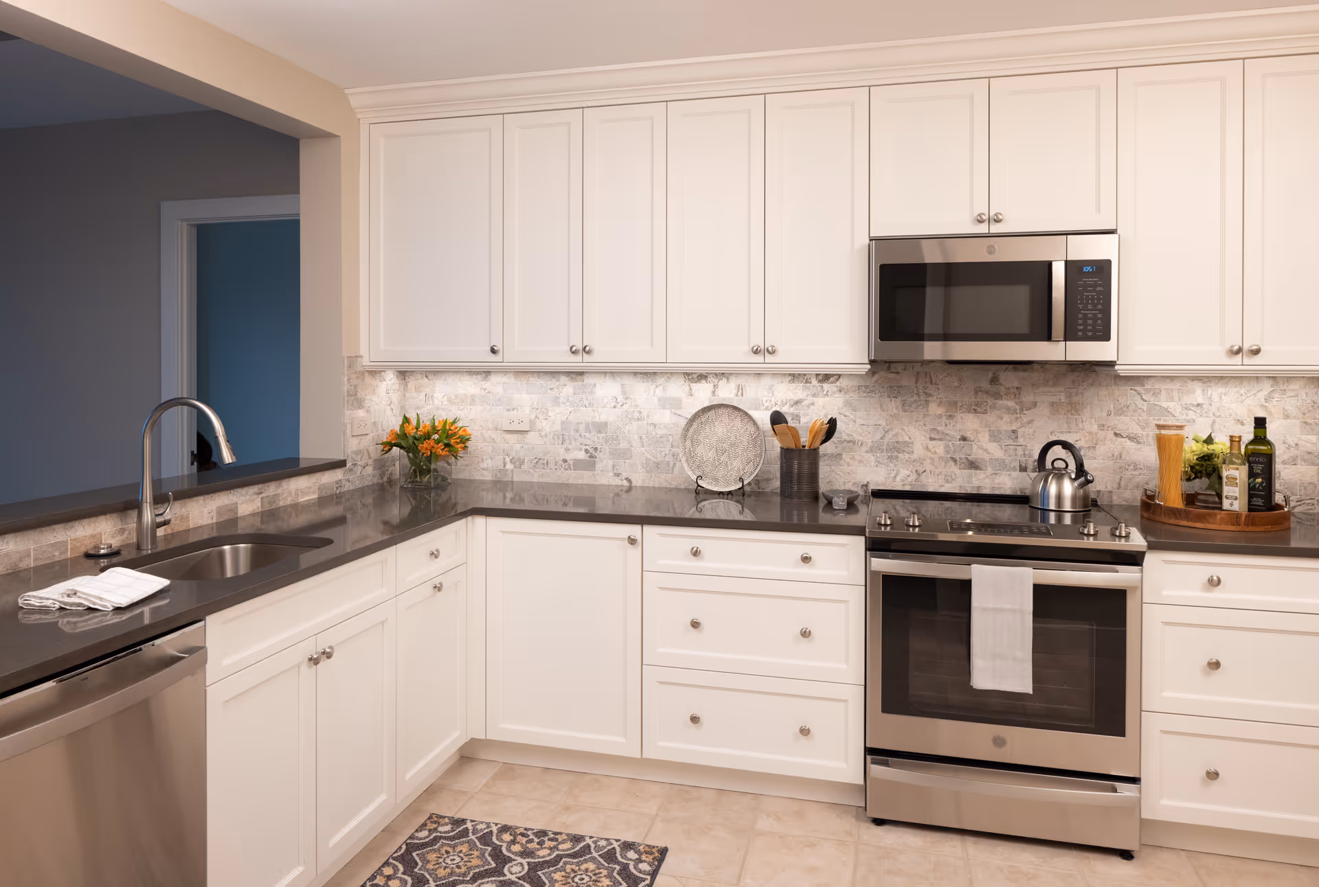 Modern kitchen with white cabinets, a stainless steel oven and microwave, a sink with a faucet, a dishwasher, and a countertop decorated with a vase of orange flowers, a decorative plate, cooking utensils, and a tray with olive oil and pasta.