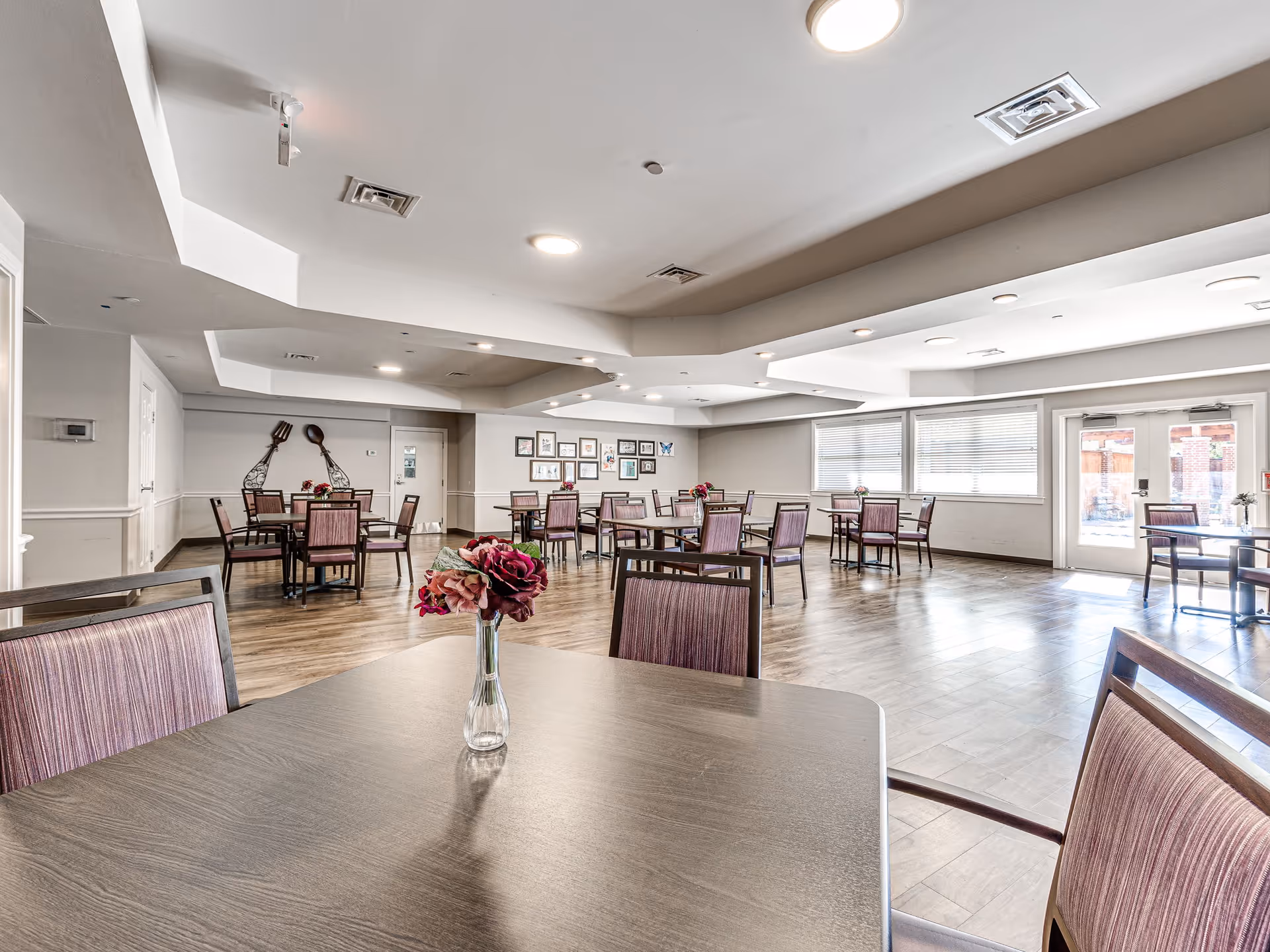 A spacious dining room with multiple tables and chairs arranged neatly. Each table has a small vase with flowers. The room has large windows allowing natural light to fill the space, light-colored walls, and a wooden floor. There are decorative oversized fork and spoon wall hangings and framed pictures on the far wall.