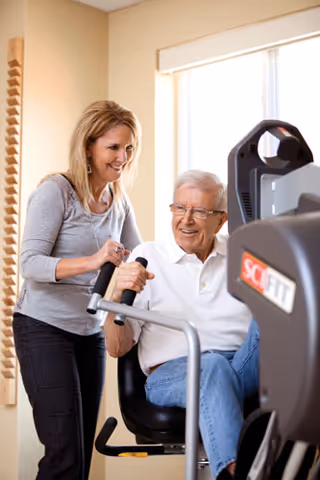 An elderly man seated on a fitness machine is assisted by a smiling woman standing beside him in a well-lit room with a window in the background.