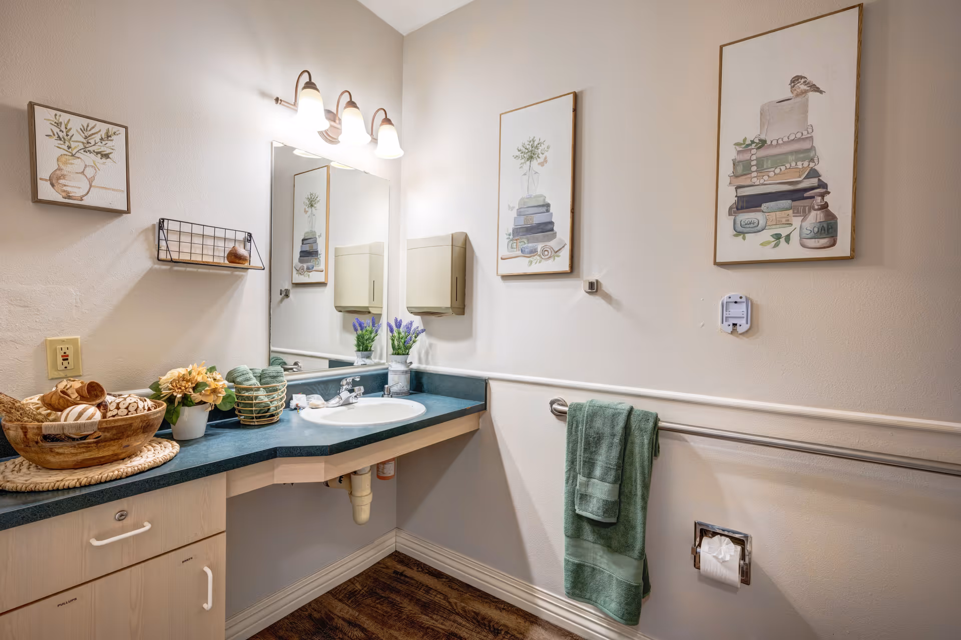 A bathroom with a green countertop and a white sink. Above the sink is a large mirror with three light fixtures. On the countertop are decorative items including a basket with towels and a small plant. The walls have framed artwork featuring books and plants. A green towel hangs on a grab bar next to a toilet paper holder. The floor is wooden.