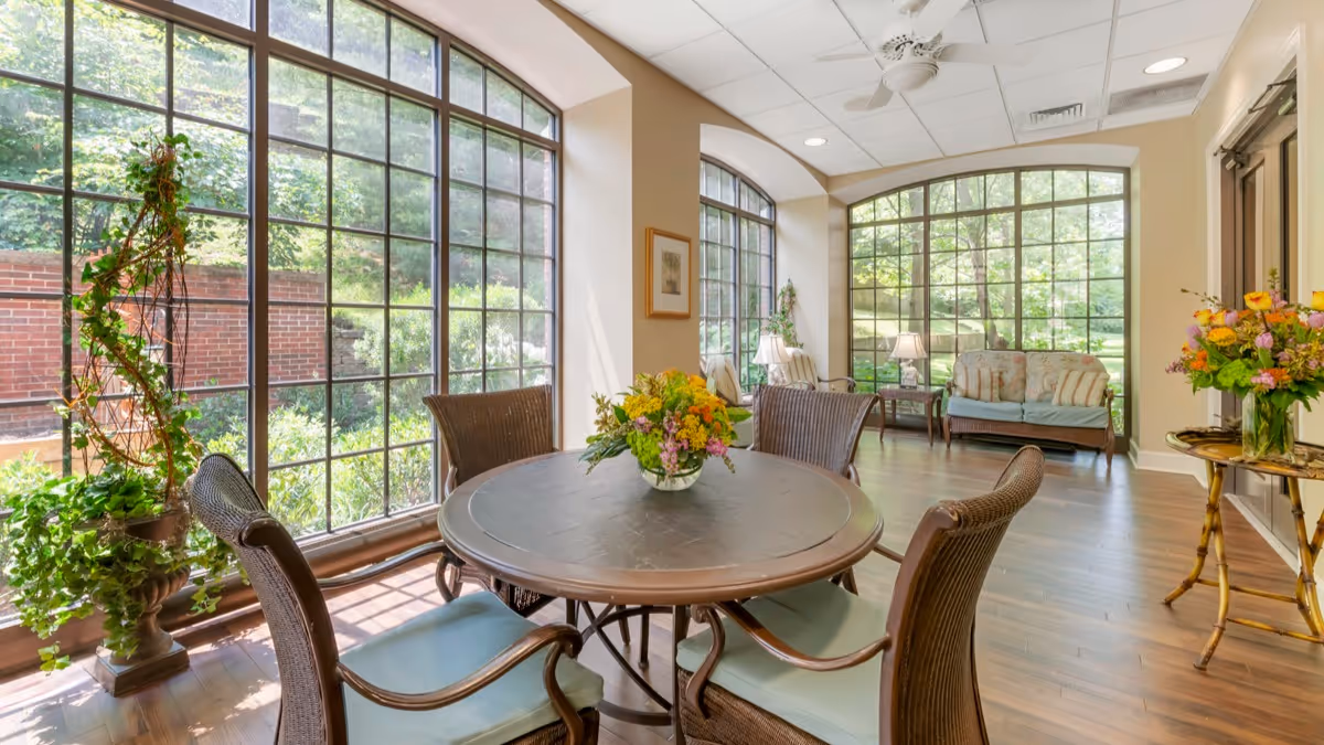 A bright and airy sitting area with large floor-to-ceiling windows overlooking a garden. The room features a round table with four wicker chairs, a vase of colorful flowers on the table, a small side table with another flower arrangement, and a cushioned bench along the far wall. The space has wooden flooring and a ceiling fan.
