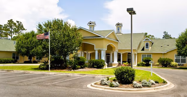 Exterior view of a single-story senior living facility building with a yellow facade, green roof, and white columns at the entrance. There is a flagpole with an American flag, landscaped bushes, and a parking lot in front under a partly cloudy sky.
