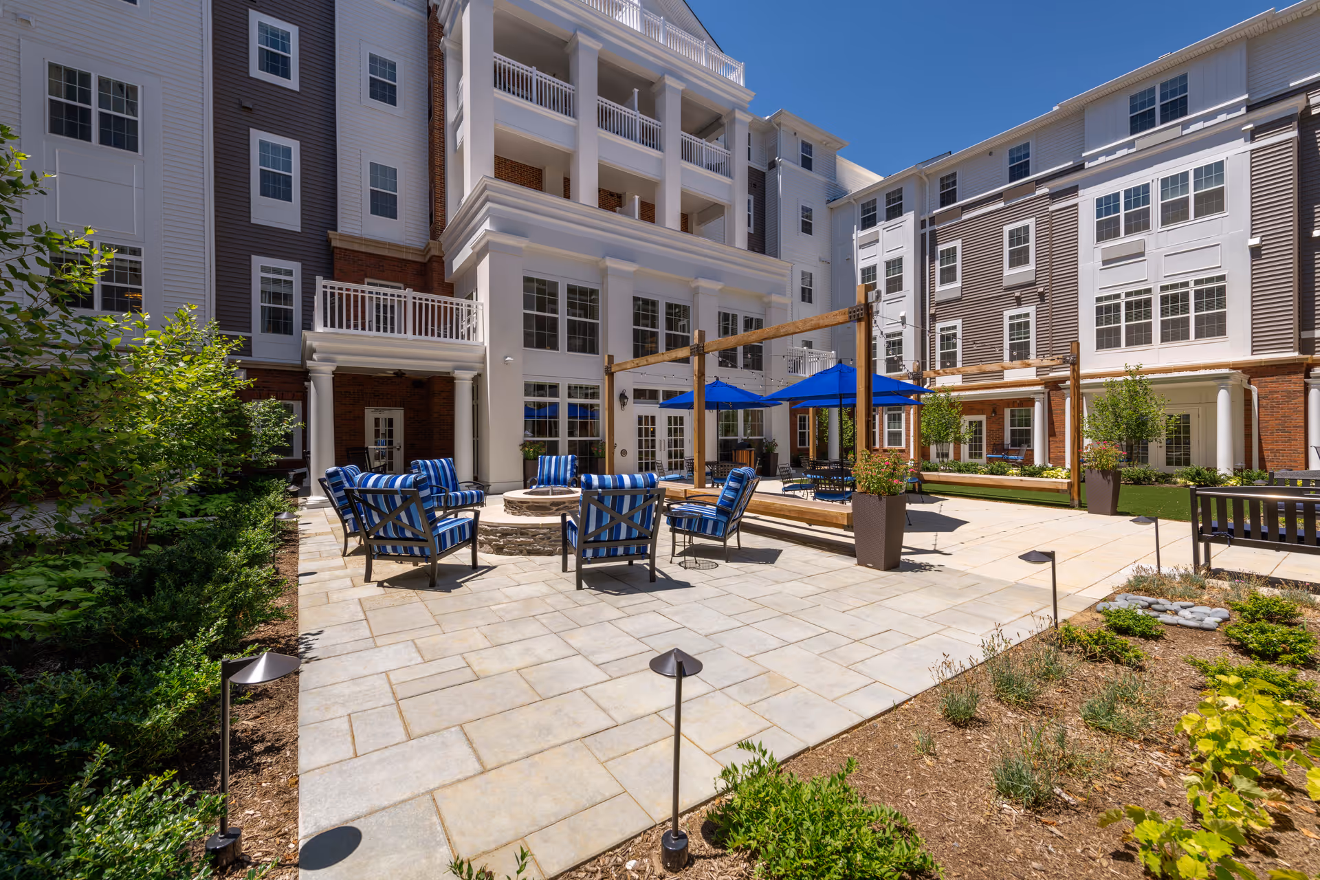 Outdoor courtyard area of a senior living facility with cushioned chairs arranged around a circular fire pit, blue umbrellas providing shade over tables, surrounded by a multi-story building with balconies and large windows, and landscaped greenery along the edges.