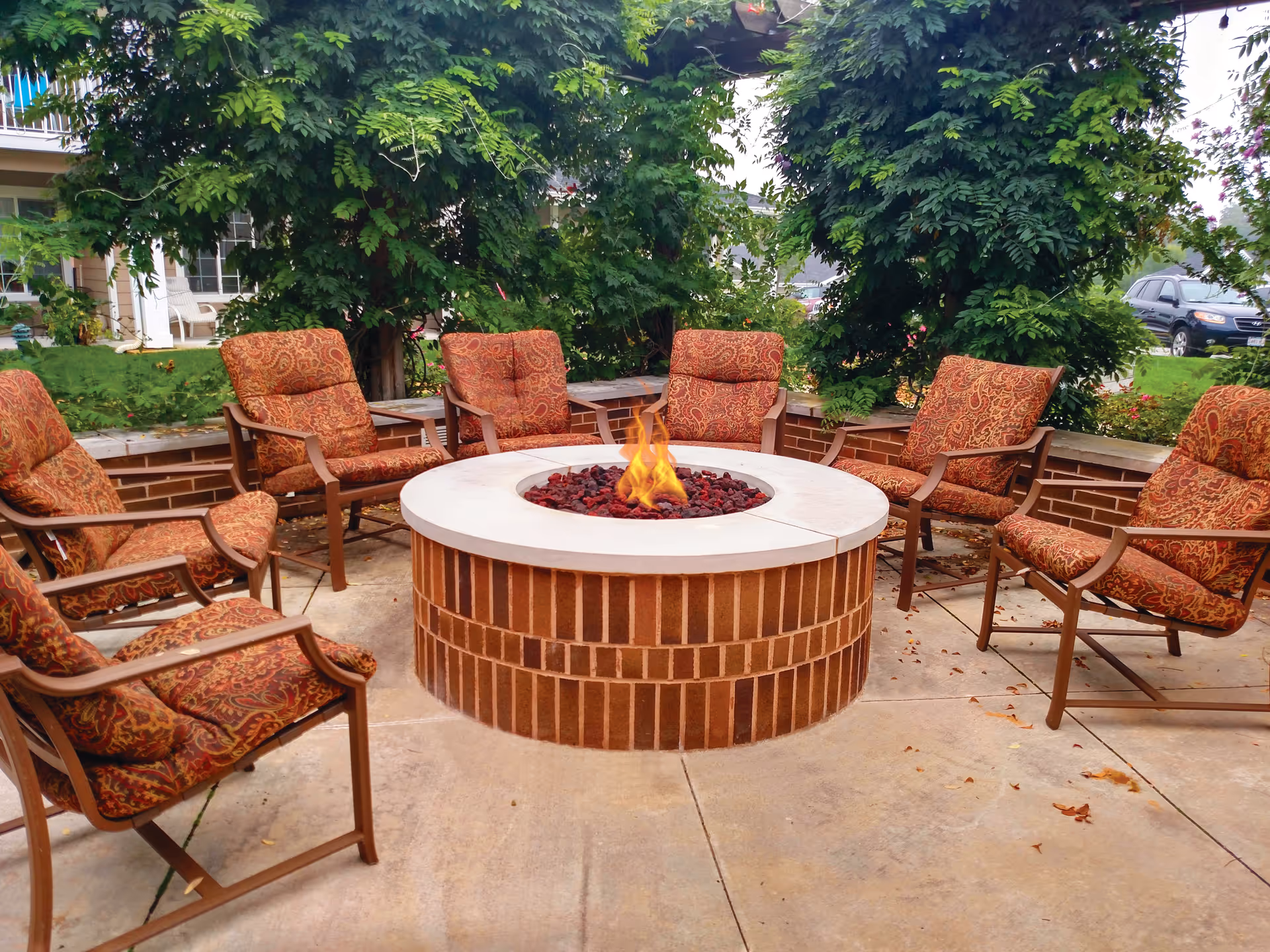 Outdoor seating area with a circular brick fire pit in the center, surrounded by eight cushioned chairs with patterned upholstery. The area is paved and bordered by greenery and trees, with a building and parked cars visible in the background.