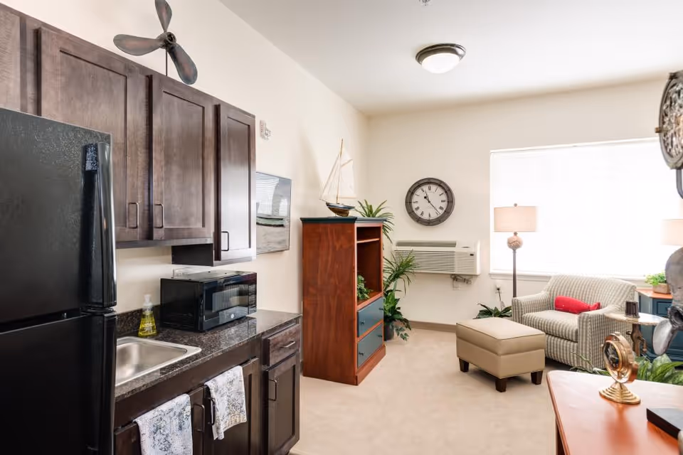 A small living area with a kitchenette featuring dark wood cabinets, a black refrigerator, a microwave, and a sink. The room also includes a beige armchair with a red pillow, a matching ottoman, a wooden shelving unit with decorative items, a wall clock, a floor lamp, and an air conditioning unit under a window with blinds.