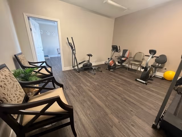 A small fitness room with exercise equipment including an elliptical machine, stationary bikes, and a yellow exercise ball. There are three cushioned chairs with wooden frames along one wall and a potted plant next to them. The room has wood-style flooring and beige walls, with an open doorway leading to another room.