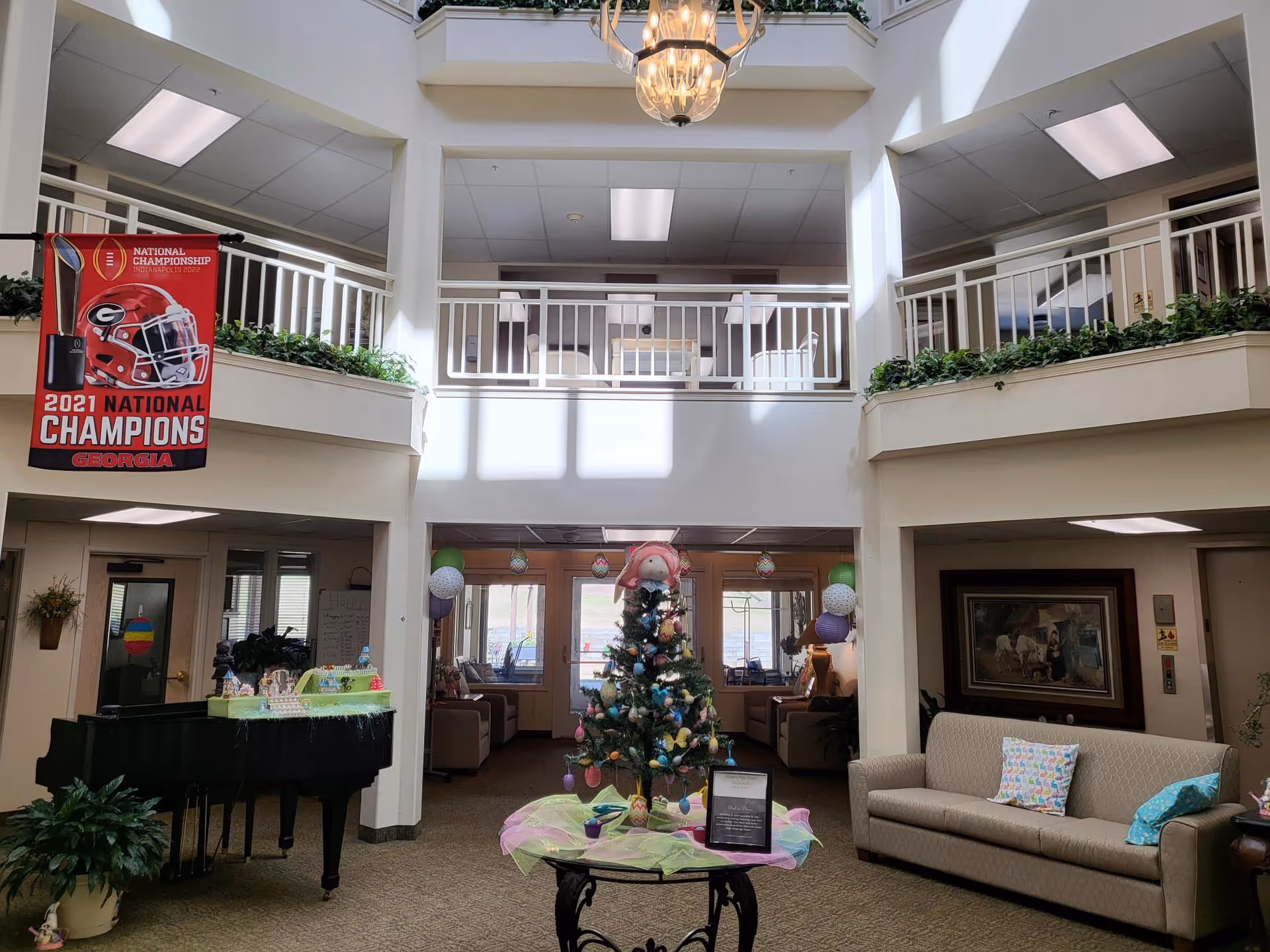Interior view of a senior living facility lobby area with a high ceiling and balcony railing on the second floor. A decorated Easter tree stands on a round table in the center, surrounded by pastel-colored decorations. To the left, there is a black grand piano with a decorative item on top. A beige couch with colorful pillows is on the right side, and a framed painting hangs on the wall behind it. A red banner celebrating the 2021 National Champions Georgia football team hangs from the balcony on the left side. The space is well-lit with natural light coming from windows in the background.