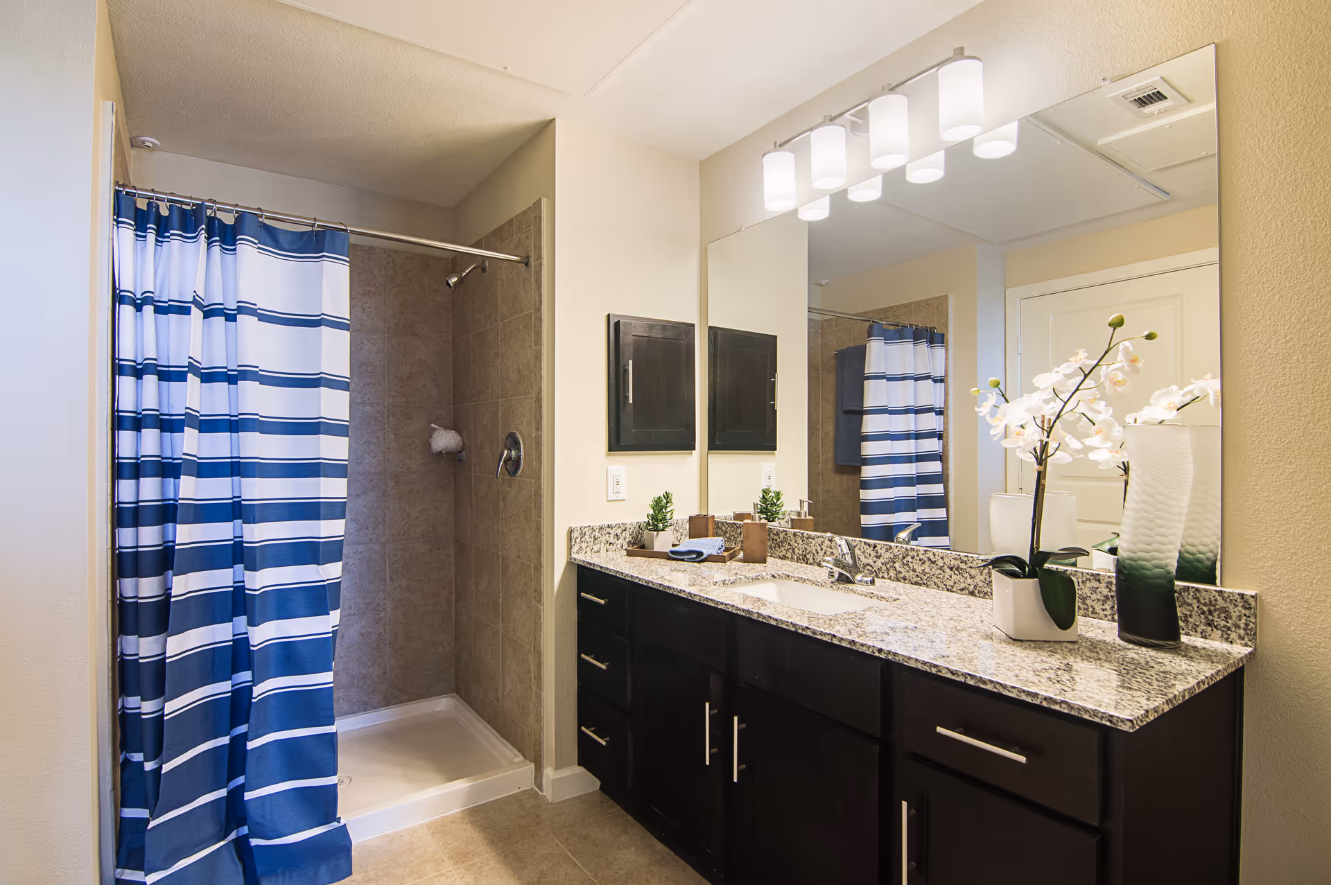 A modern bathroom featuring a walk-in shower with a blue and white striped shower curtain, a granite countertop with a sink, dark wood cabinets, a large mirror, and decorative plants including white orchids.