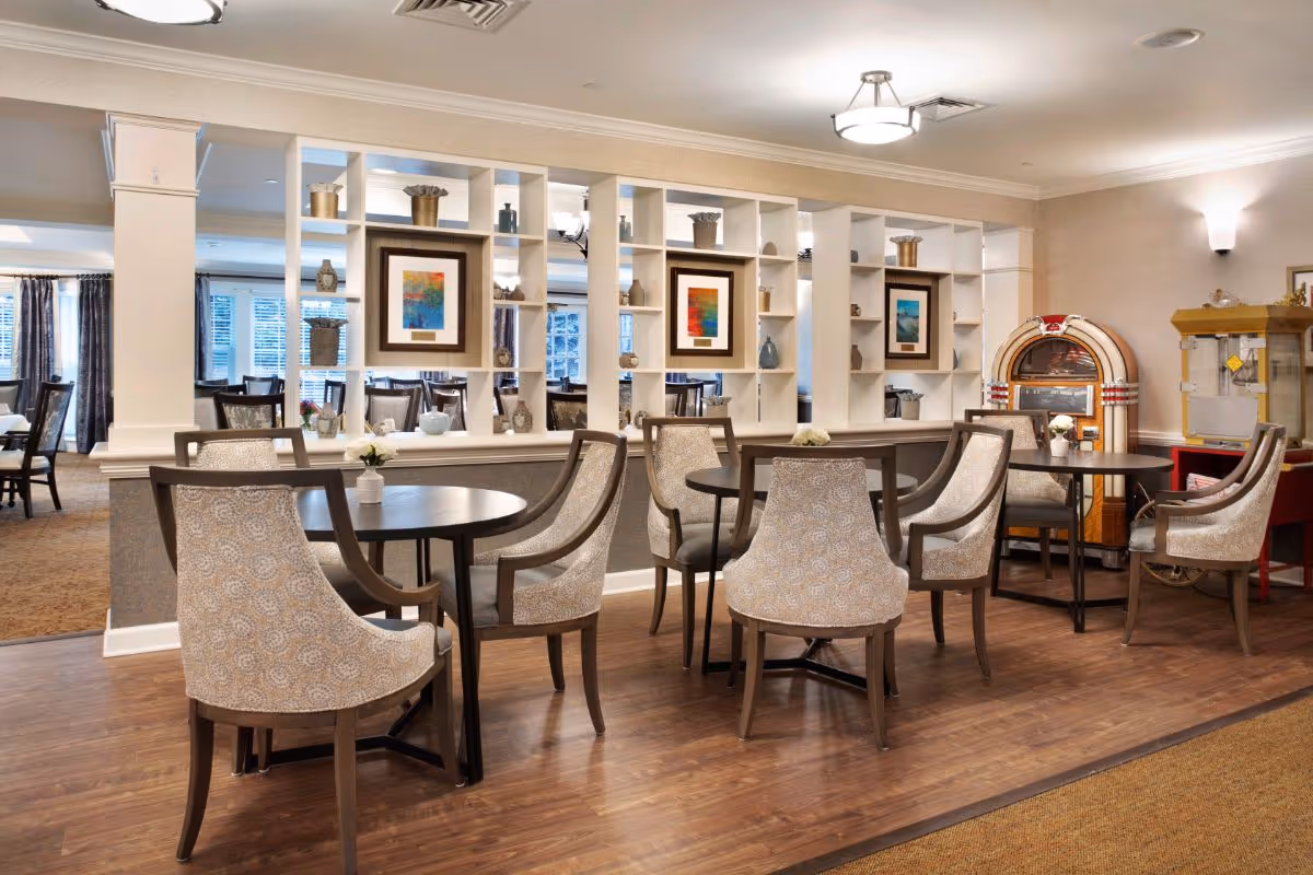 A dining area in a senior living facility with round tables and upholstered chairs arranged neatly. The room features a decorative white shelving divider with framed colorful artwork and various decorative items. In the background, there is a vintage-style jukebox and a popcorn machine. The space has warm lighting and wood flooring.