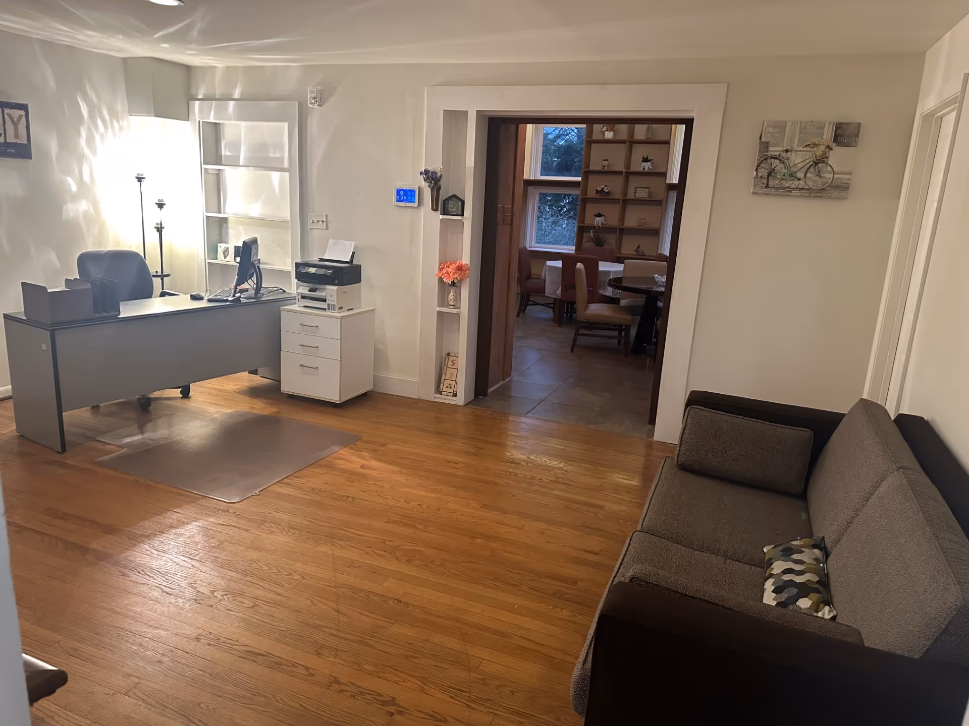Interior view of a room with wooden flooring featuring a gray office desk with a computer and printer on the left side, a gray couch with two cushions on the right side, and an open doorway leading to a dining area with a round table and chairs. The walls are white with minimal decorations including a bicycle painting and small shelves with flowers.