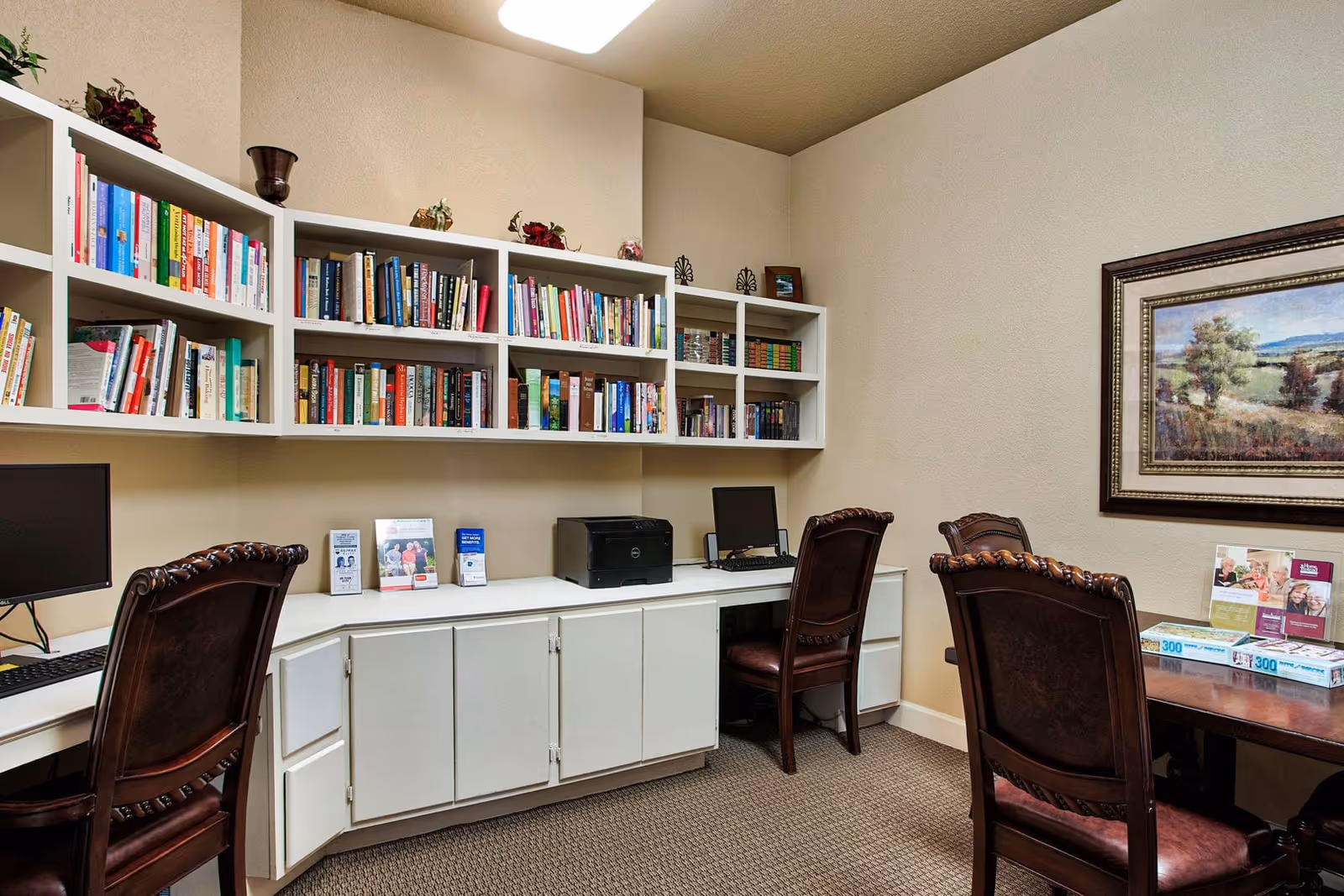 A cozy room with a built-in white bookshelf filled with books, two computer workstations with monitors and keyboards, a printer on the counter, and dark wooden chairs. There is a framed landscape painting on the wall and a table with brochures and puzzle boxes.