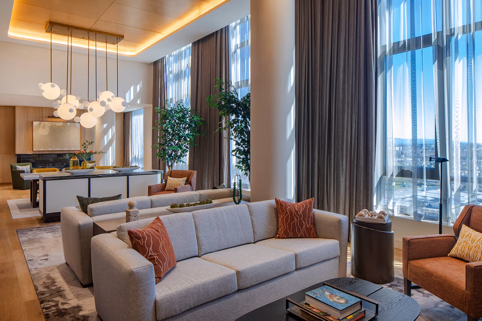 A modern living room with large floor-to-ceiling windows covered by sheer curtains, allowing natural light to fill the space. The room features a light gray sofa with rust-colored pillows, an orange armchair with a yellow pillow, a dark coffee table with books, and a side table with decorative items. In the background, there is a dining area with a long table, chairs, pendant lights, and two tall green plants near the windows.
