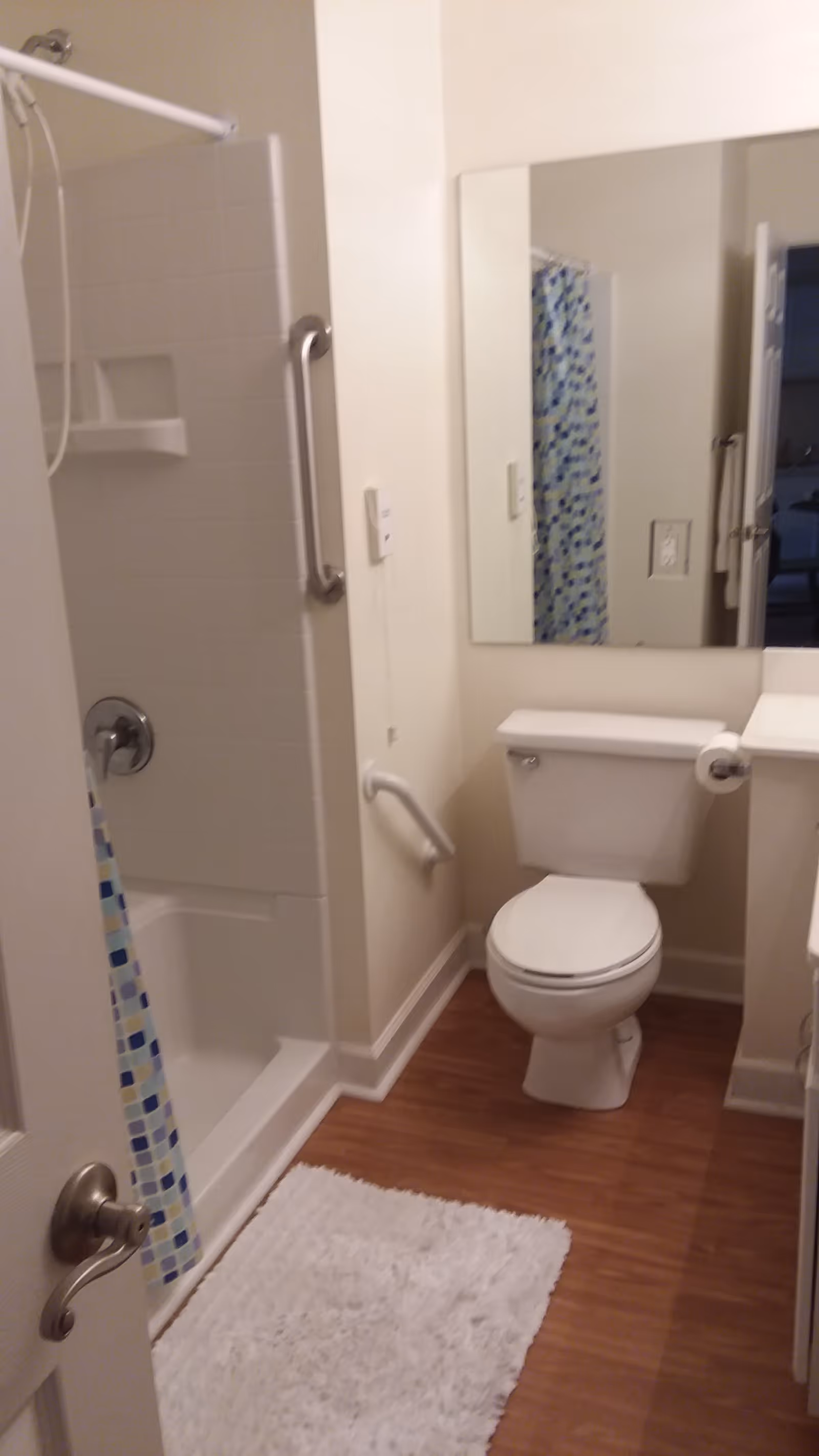 A small bathroom with a white toilet, a white shower with a blue and white patterned shower curtain, a large mirror above the toilet, a white rug on the wooden floor, and grab bars installed near the shower and toilet for accessibility.