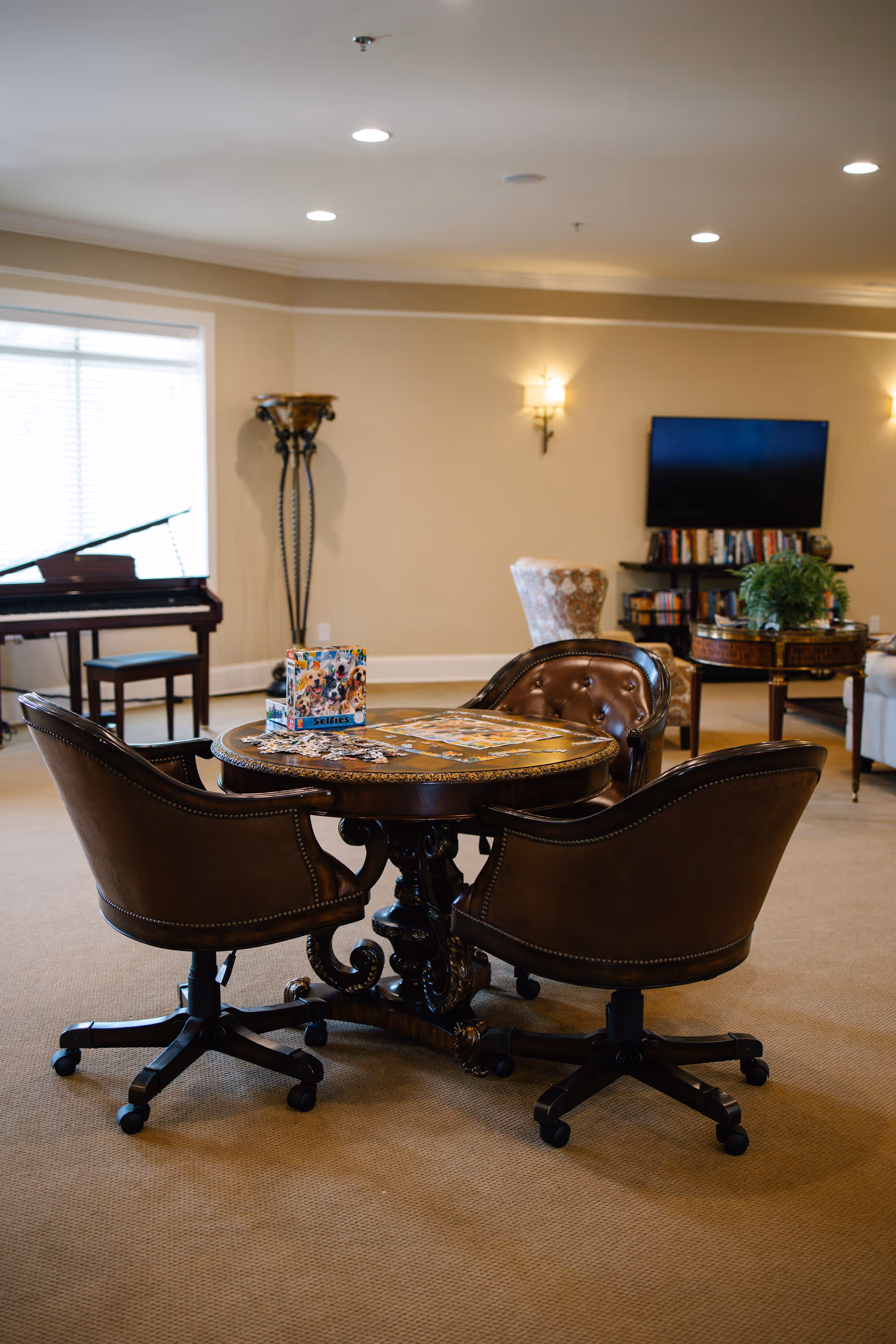 A cozy living room area with a round wooden table surrounded by four brown leather swivel chairs. On the table, there is a puzzle and a box labeled 'Selfies.' In the background, there is a piano near a window, a tall decorative lamp, a TV mounted on the wall with a bookshelf underneath, and a small round table with a plant on it. The room has beige walls and carpeted floor with recessed ceiling lights.