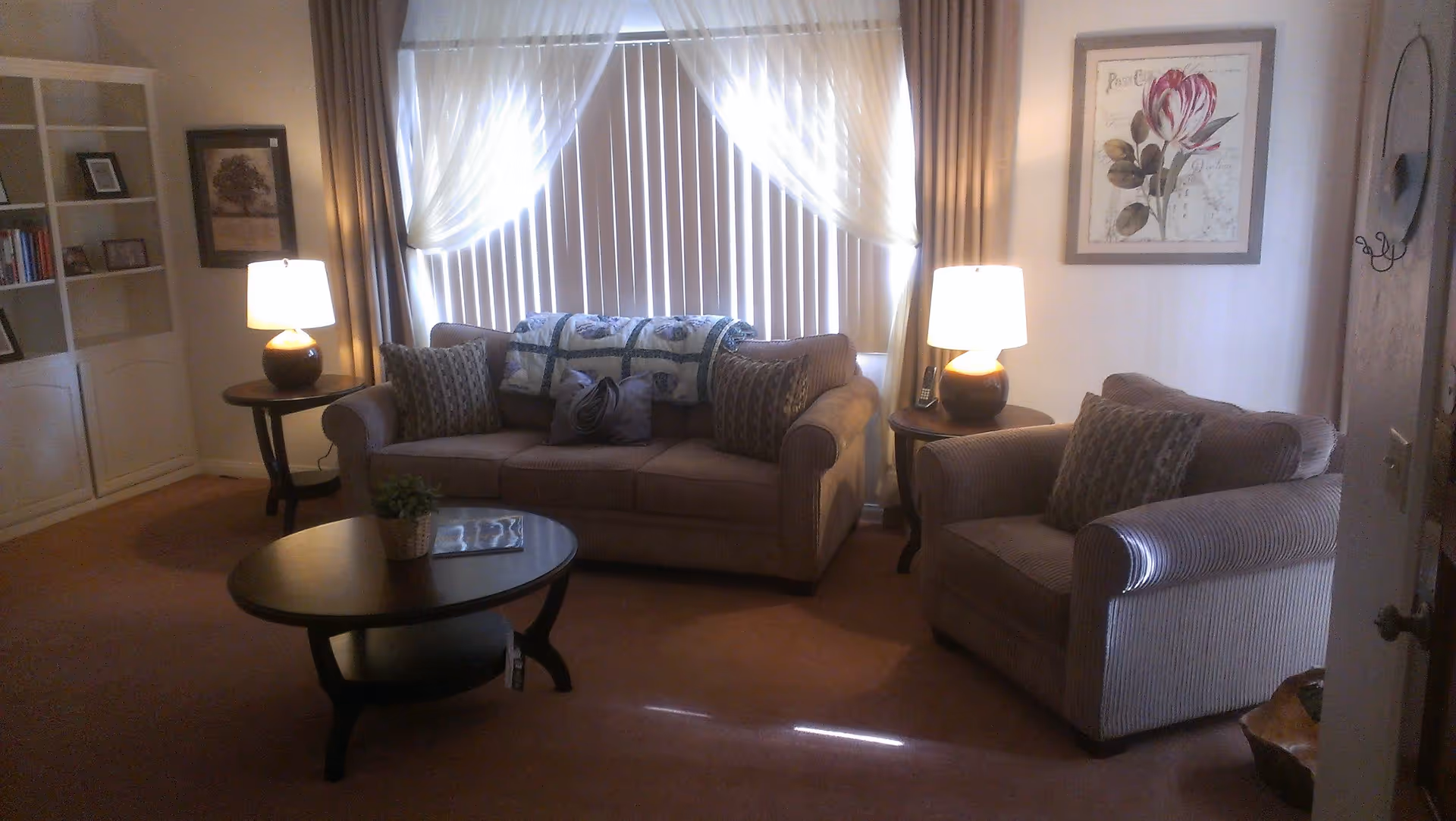 A cozy living room with a brown sofa and matching armchair, both adorned with patterned pillows. Two side tables with lamps flank the sofa, and a round coffee table with a small plant and a magazine sits in front. The window behind the sofa is covered with vertical blinds and sheer curtains. The walls feature framed artwork and a round mirror with hooks.