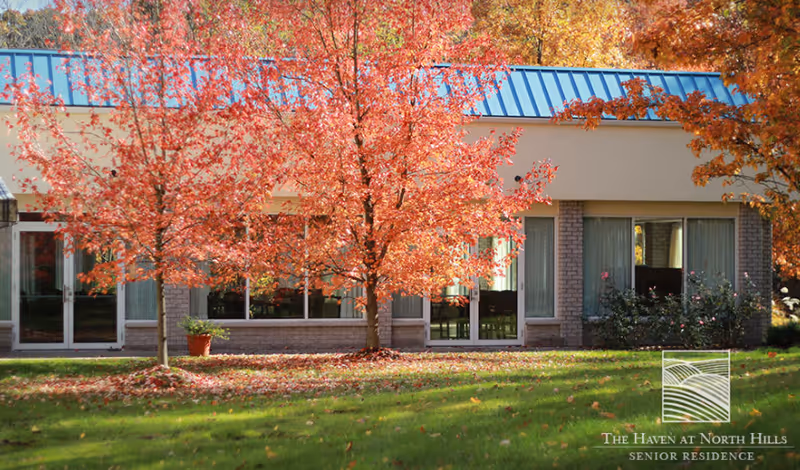 Exterior view of a senior living facility building with large windows and a blue roof, surrounded by green grass and trees with vibrant red and orange autumn leaves.