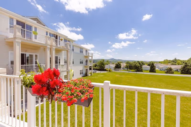 View from a balcony with white railing adorned with red flowers, overlooking a large green lawn and a multi-story residential building under a blue sky with scattered clouds.