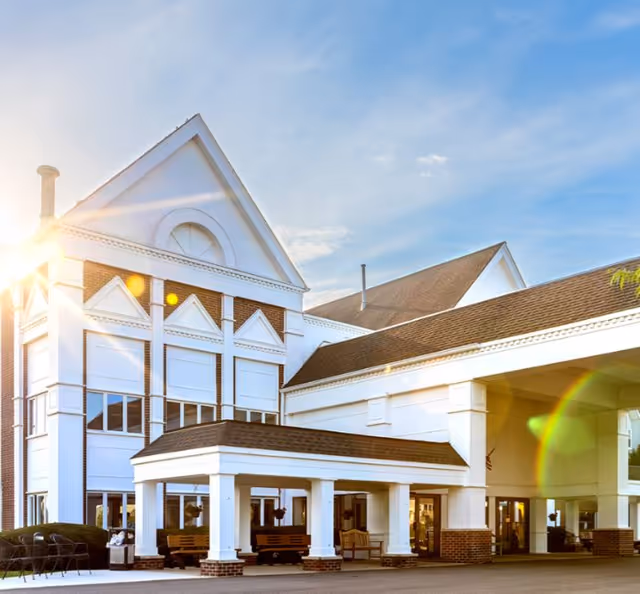 Exterior view of a large, white senior living facility building with brick accents under a clear blue sky with the sun shining brightly, featuring a covered entrance with benches and large windows.