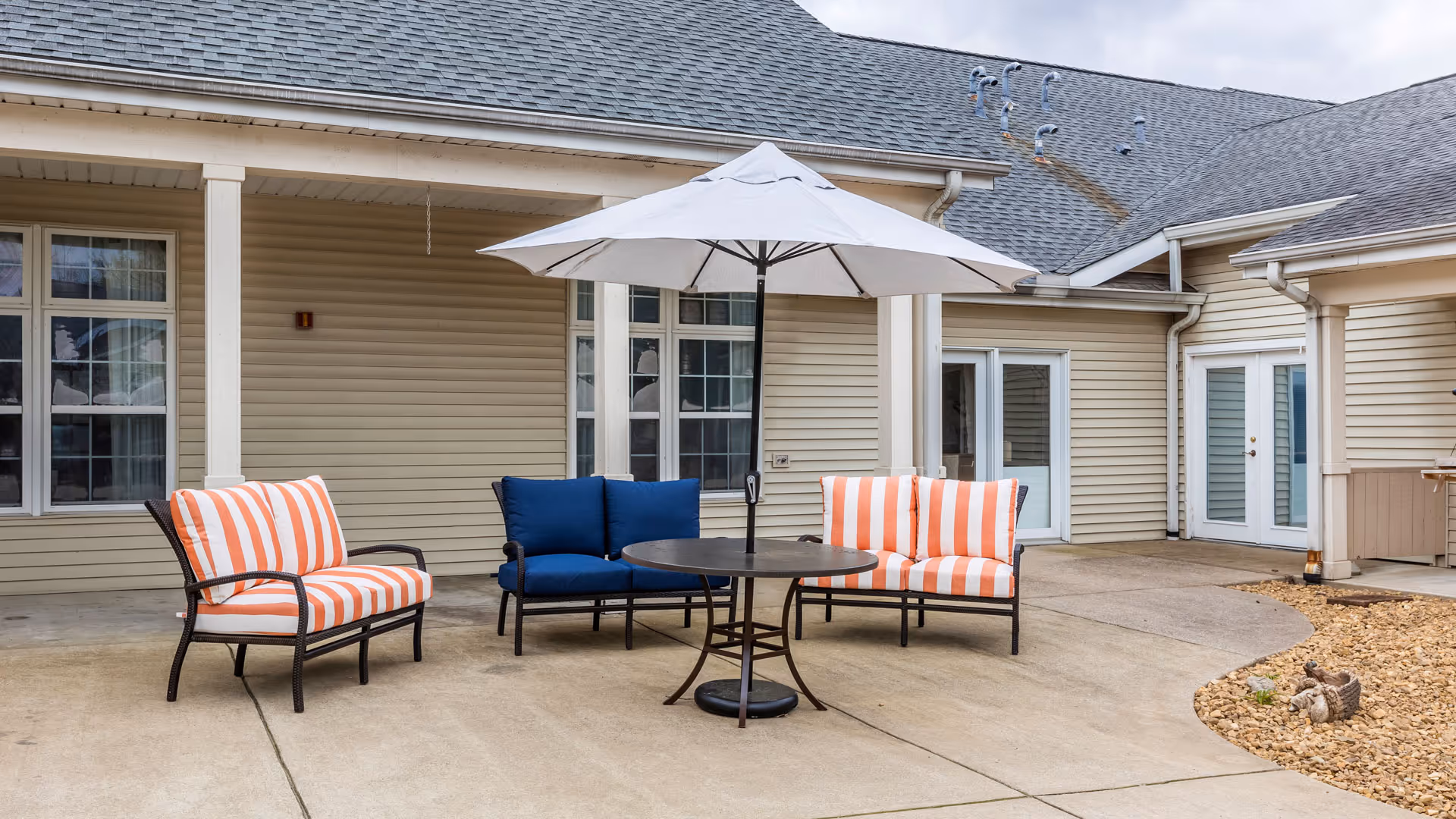 Patio with orange-and-white striped chairs, a blue loveseat, round table and umbrella in front of a beige building.