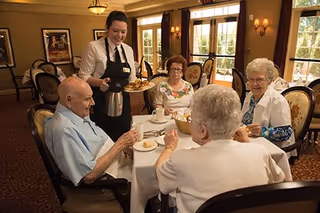 A waitress serving coffee and food to four elderly people seated around a dining table in a well-lit dining room with large windows and elegant decor.